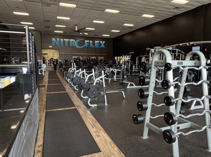 Spacious gym interior with rows of exercise bikes and weight machines, black walls, and a NITROFLEX sign.