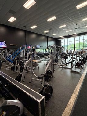 Busy gym interior with weight machines, treadmills, mirrors, and overhead fluorescent lights.