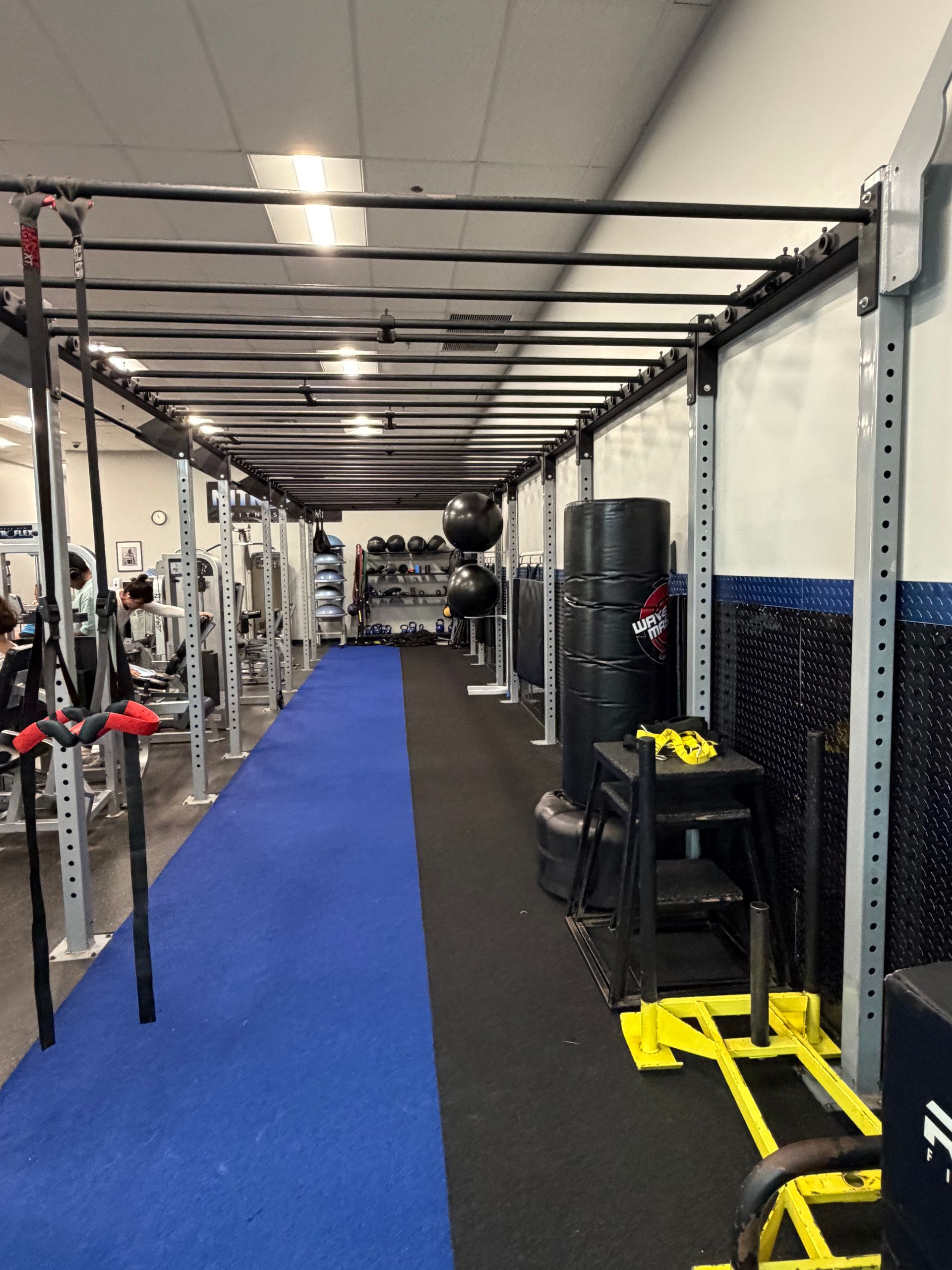Gym interior with a blue turf lane, weight racks, and exercise equipment along a narrow training area