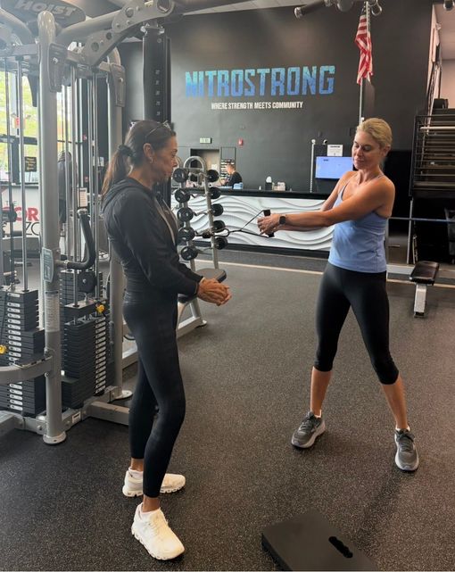 Two women exercising with a cable machine in a gym, one demonstrating a movement to the other.