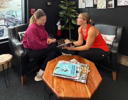 Two people talking at a table in a gym, one writing in a notebook while the other listens.
