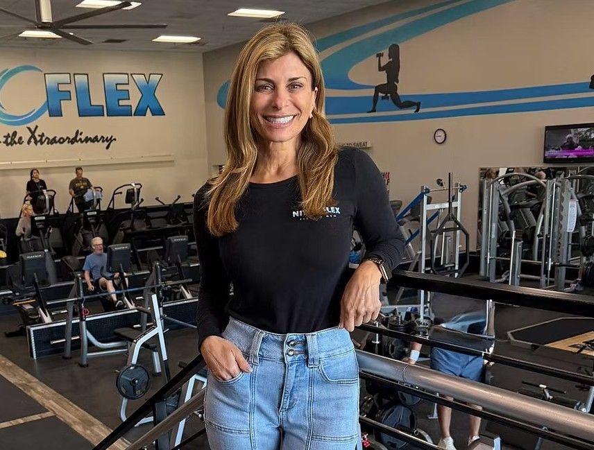 Woman standing in a gym, smiling in a black top and light blue jeans.
