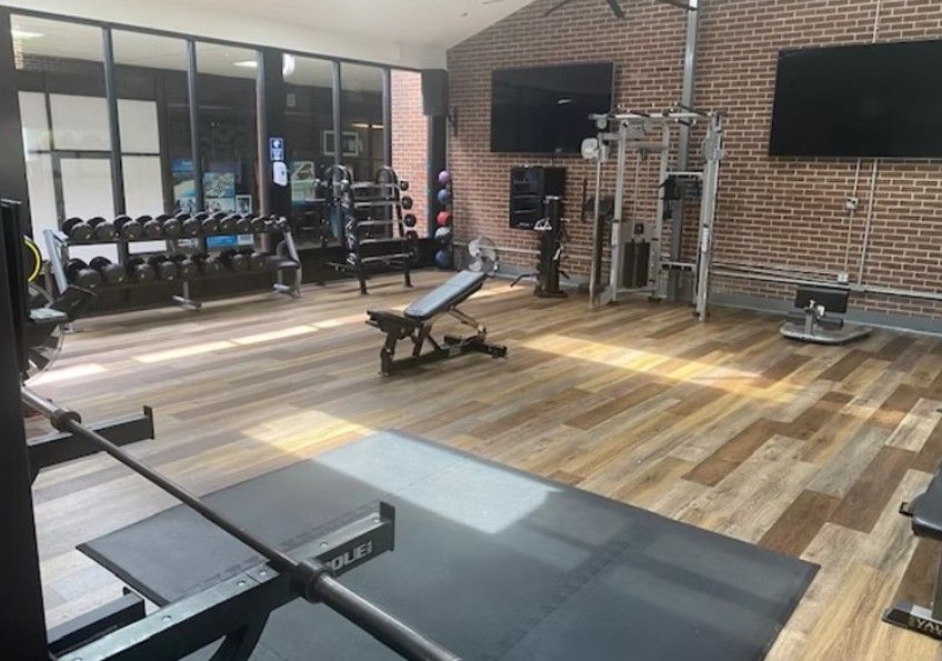 Modern gym with wood floors, weight benches, dumbbells, and TVs against brick and glass walls
