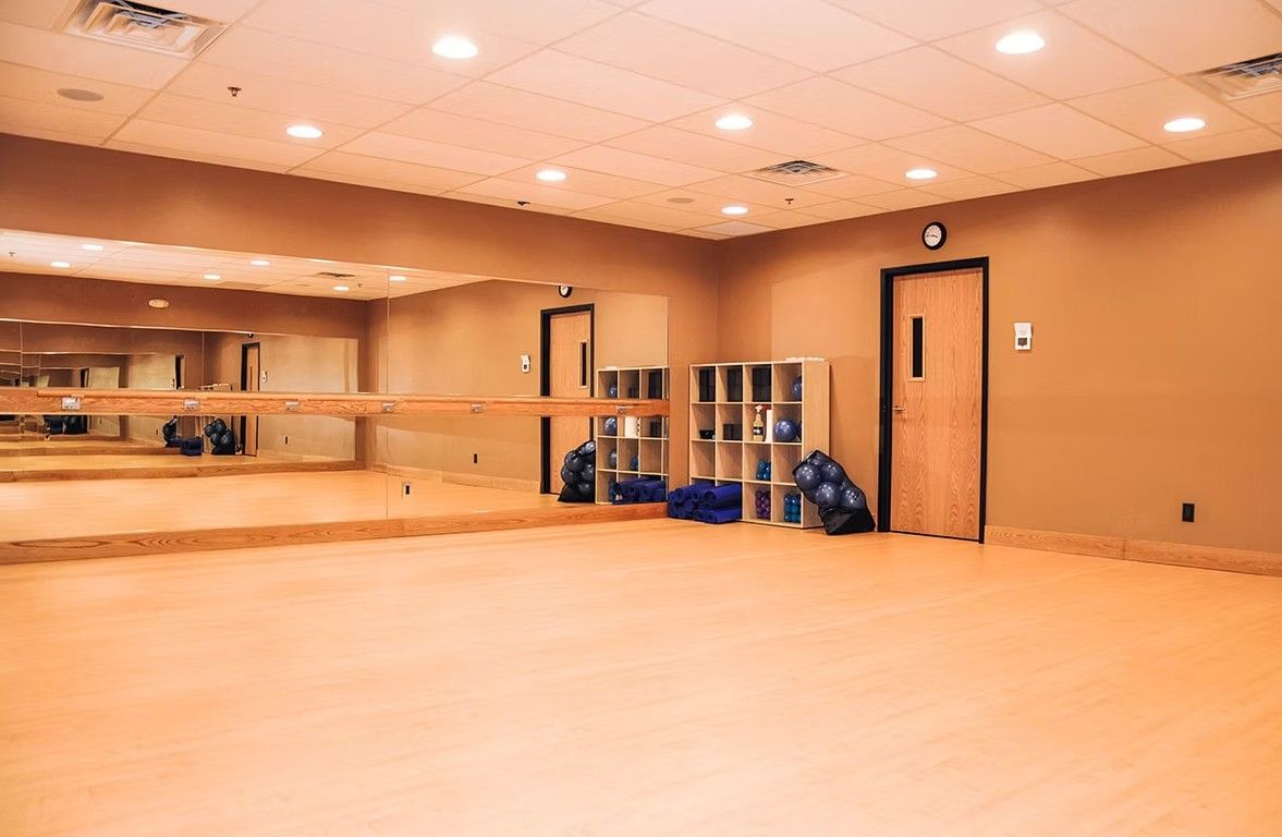 Empty dance studio with mirrored wall, wood floor, ballet barres, and bags near the door.