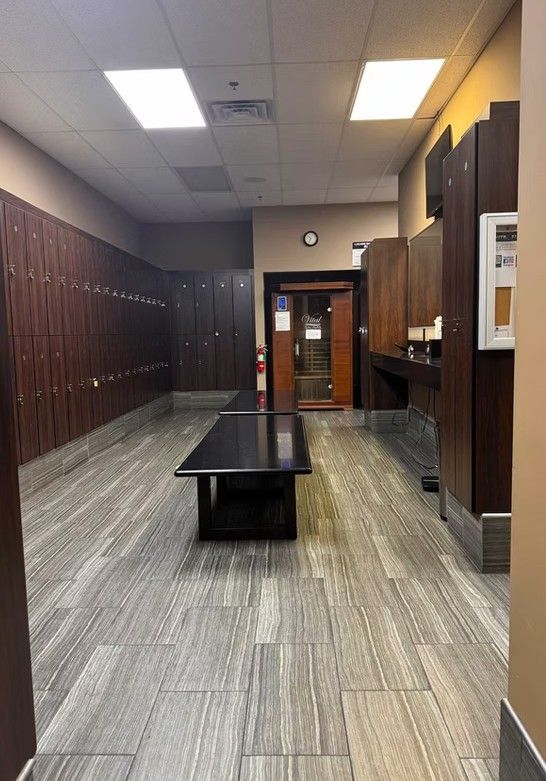 Empty locker room with brown lockers, gray floor, and central black bench under fluorescent lights