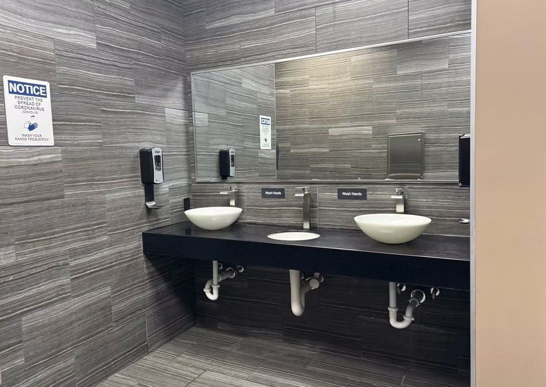 Public restroom with two white sinks, black counter, and gray tiled walls.