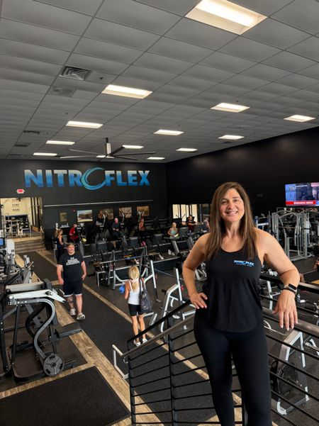 Nitroflex gym interior with a smiling woman posing by stairs and workout equipment.