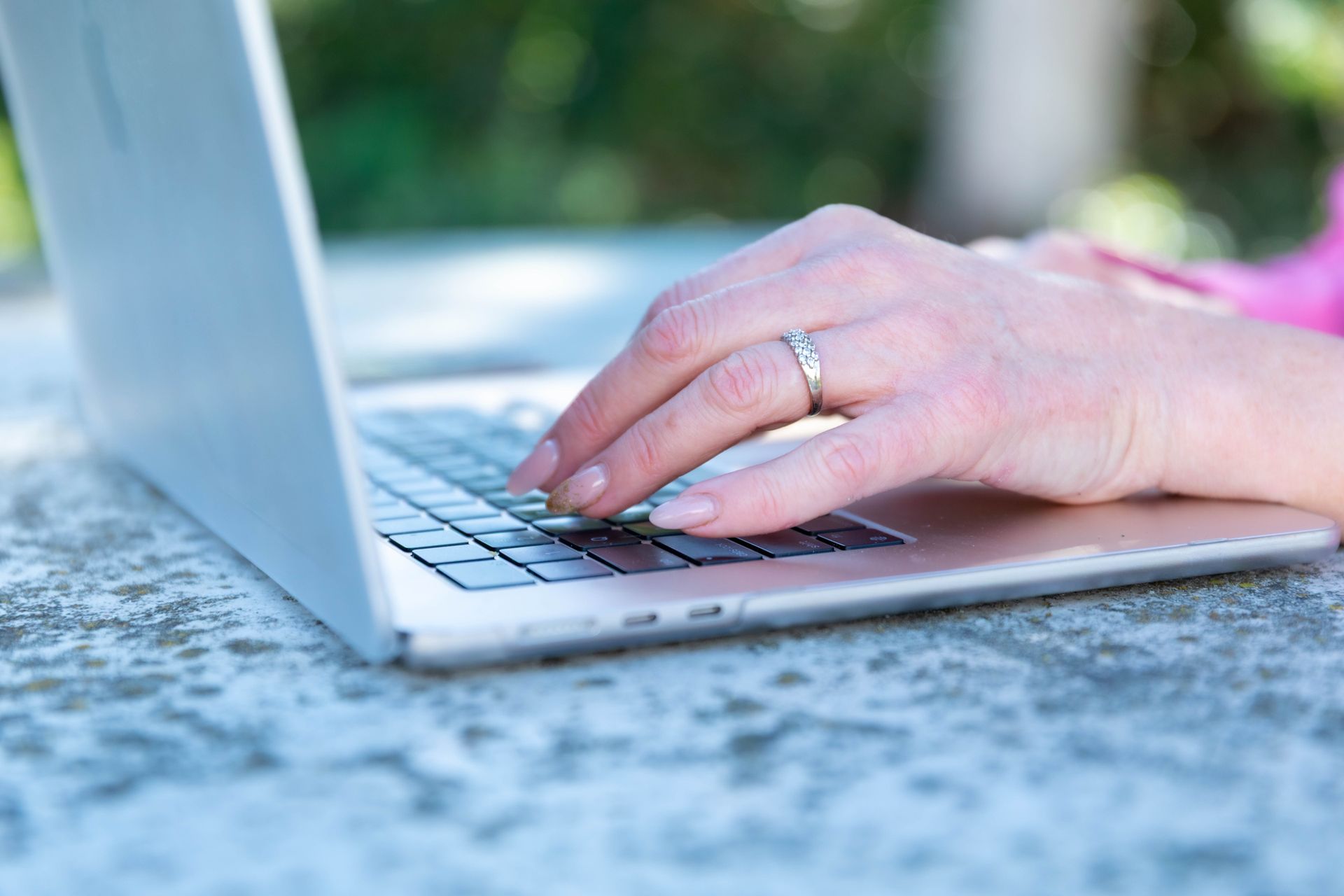 Hands typing on a silver laptop outdoors on a stone table.