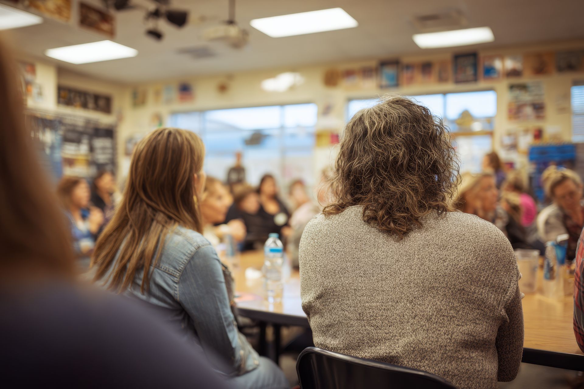 People seated at a classroom table listening to a group discussion in a bright room.