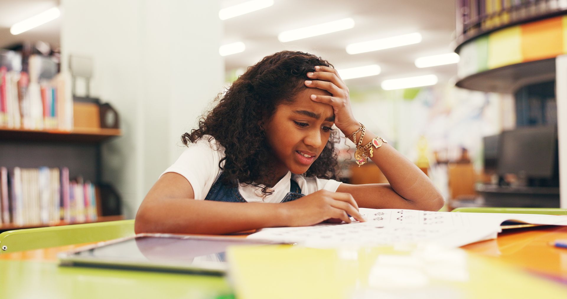 Student studying at a table in a library, leaning over papers with a focused expression.