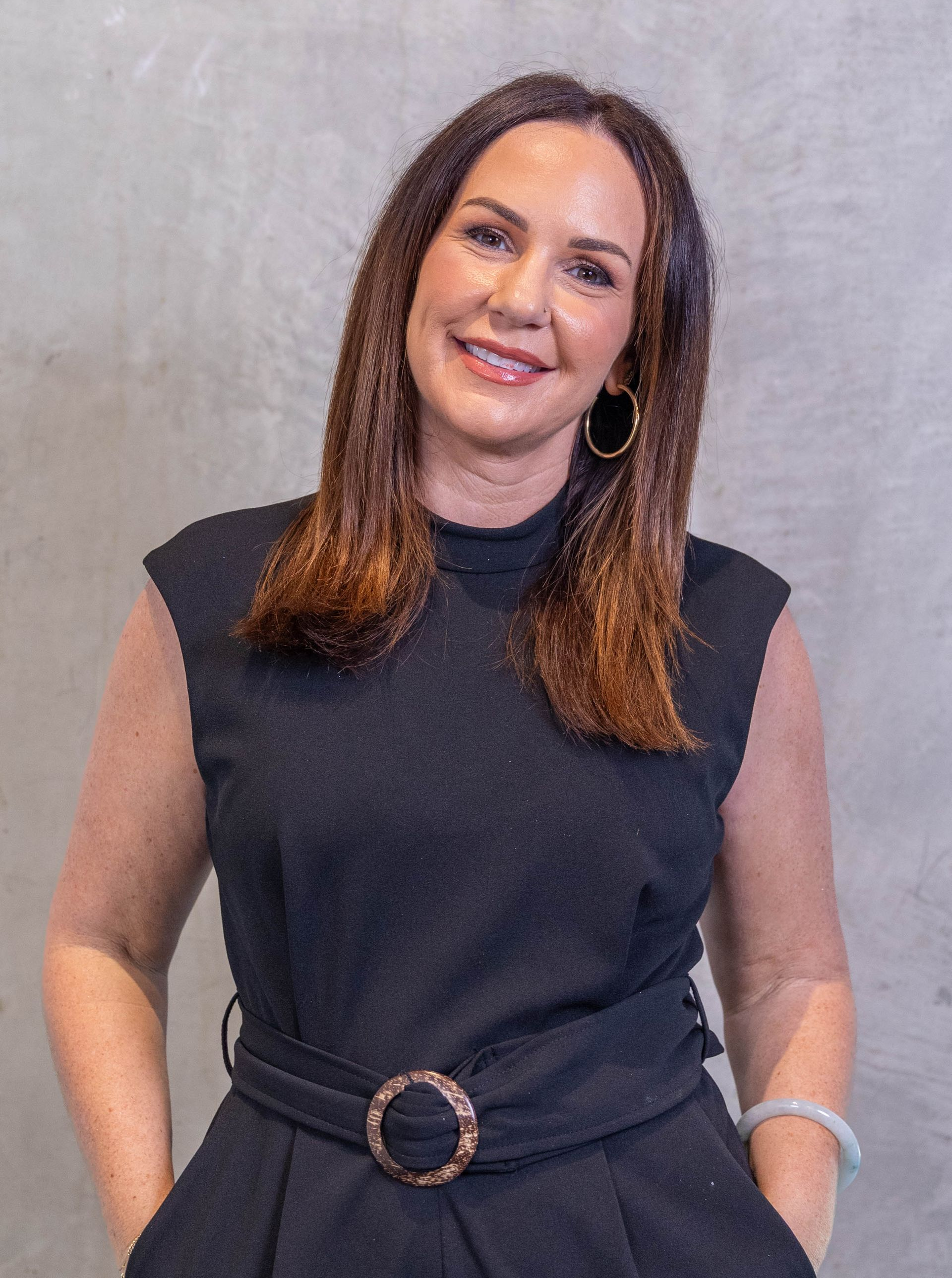 Woman in a sleeveless black dress with a belt, smiling against a gray backdrop.