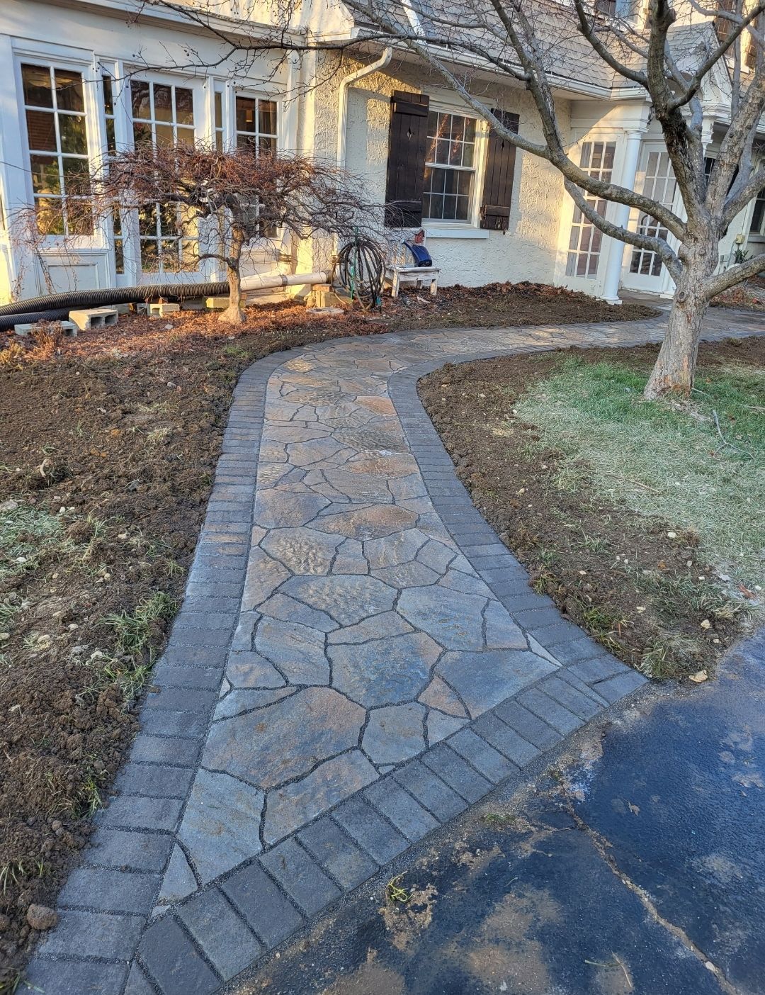 Stone walkway with brick border leading to a house, with bare landscaping on either side.