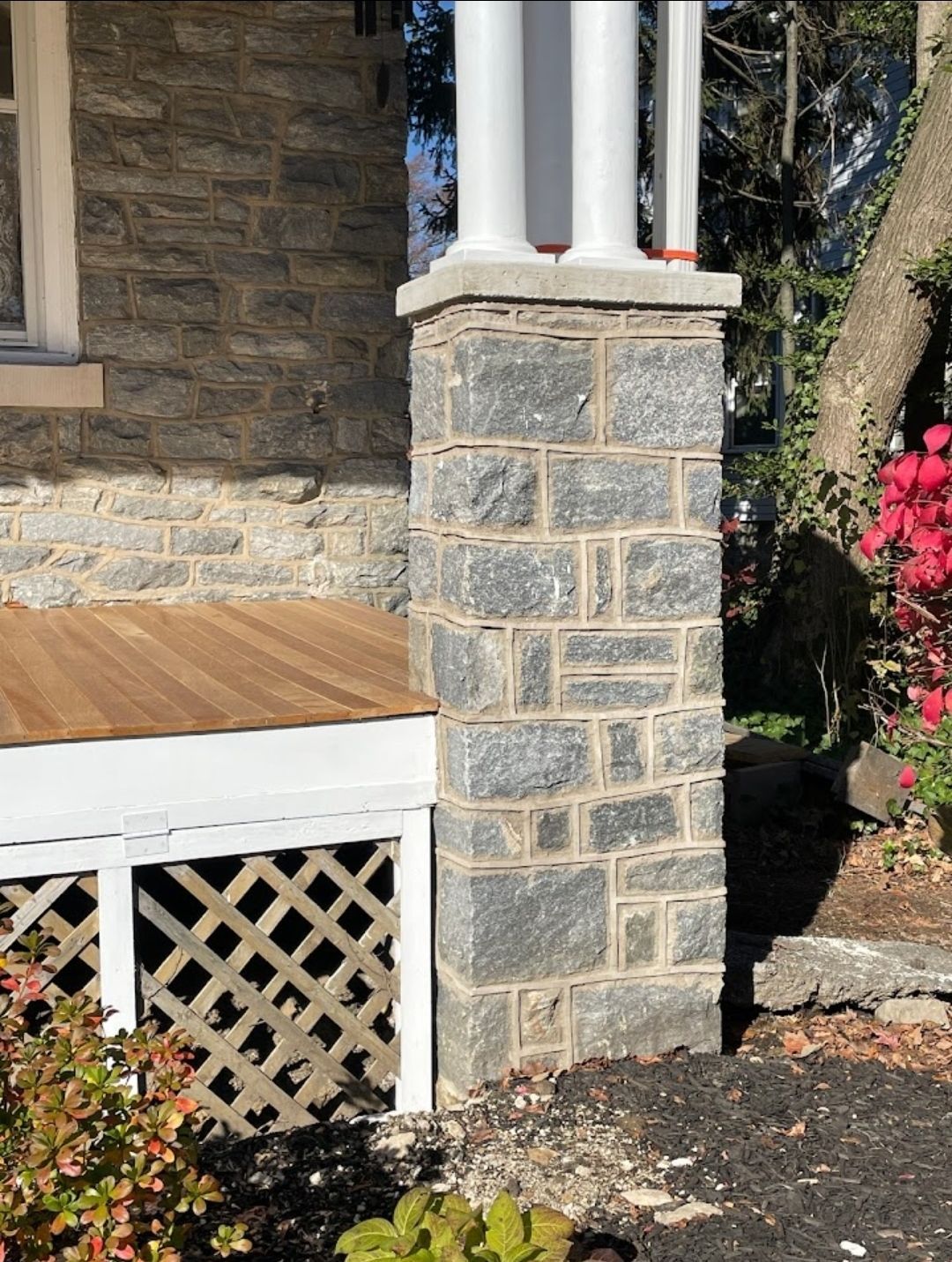 Stone column supporting a white-pillared roof. Lattice detail underneath. Foliage surrounds.