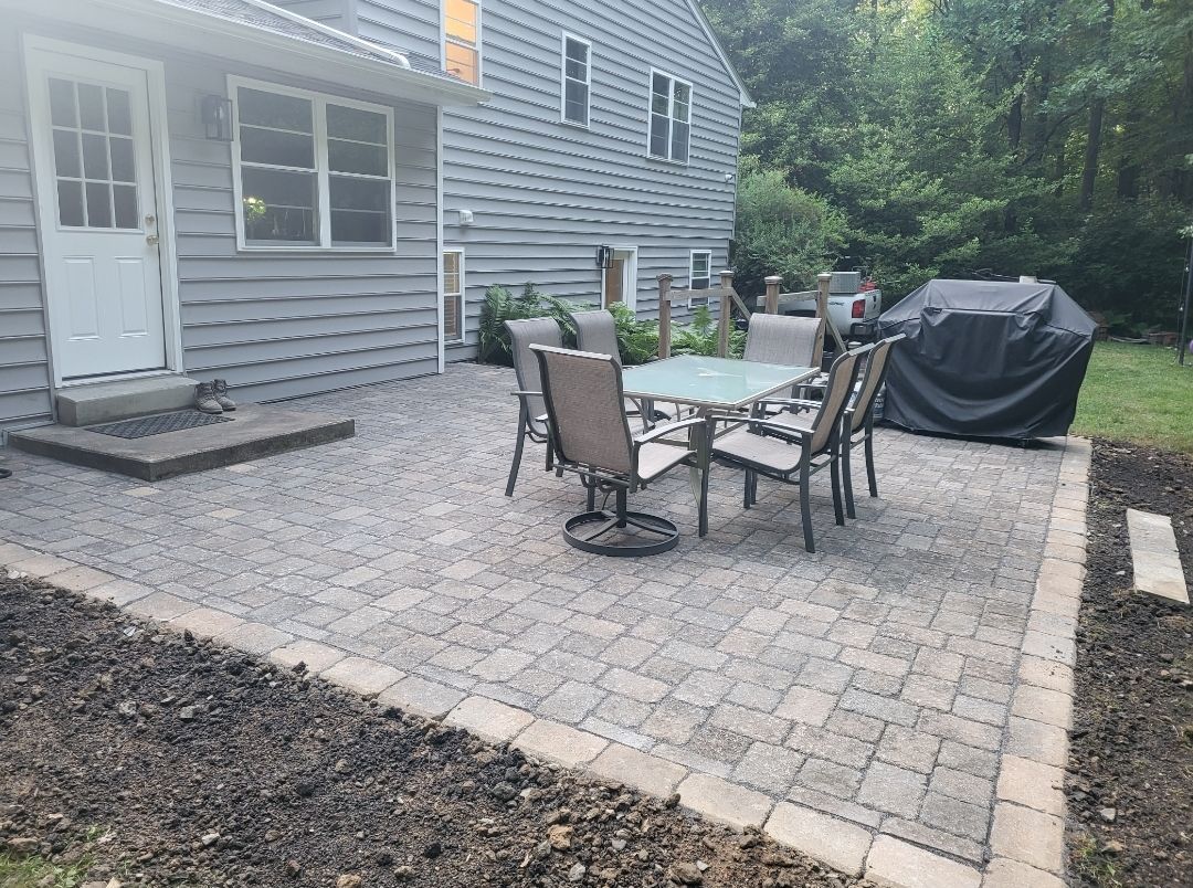Patio with table, chairs, and grill next to a gray house, surrounded by dirt and grass.