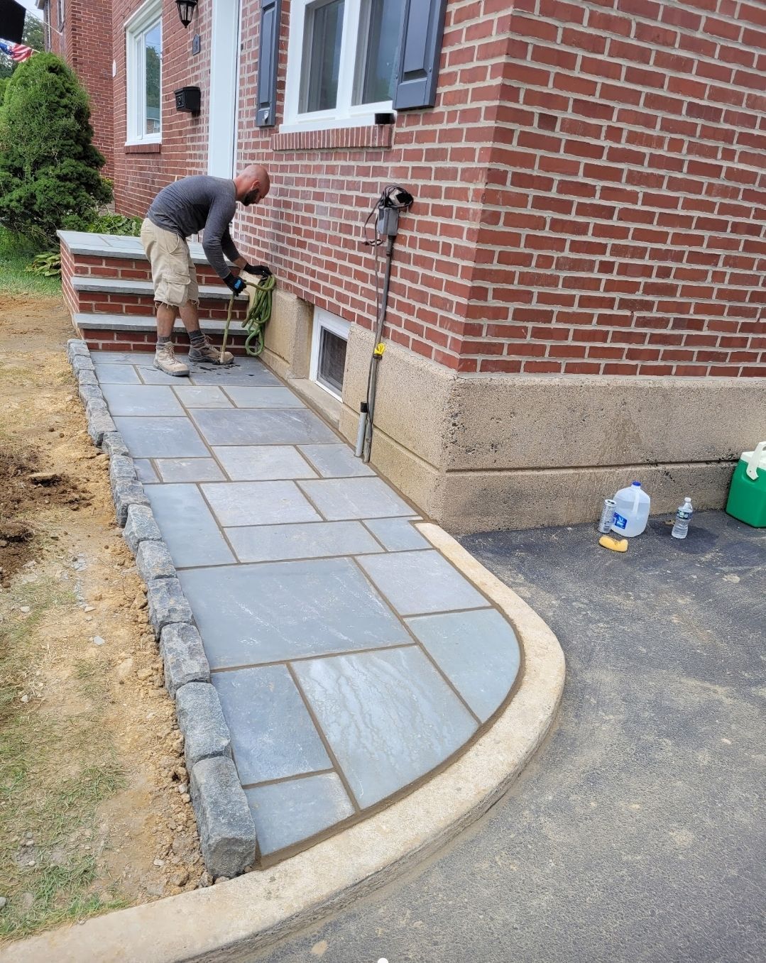 A person installing a stone walkway alongside a red brick house; dark gray pavers and a curved light-brown border.