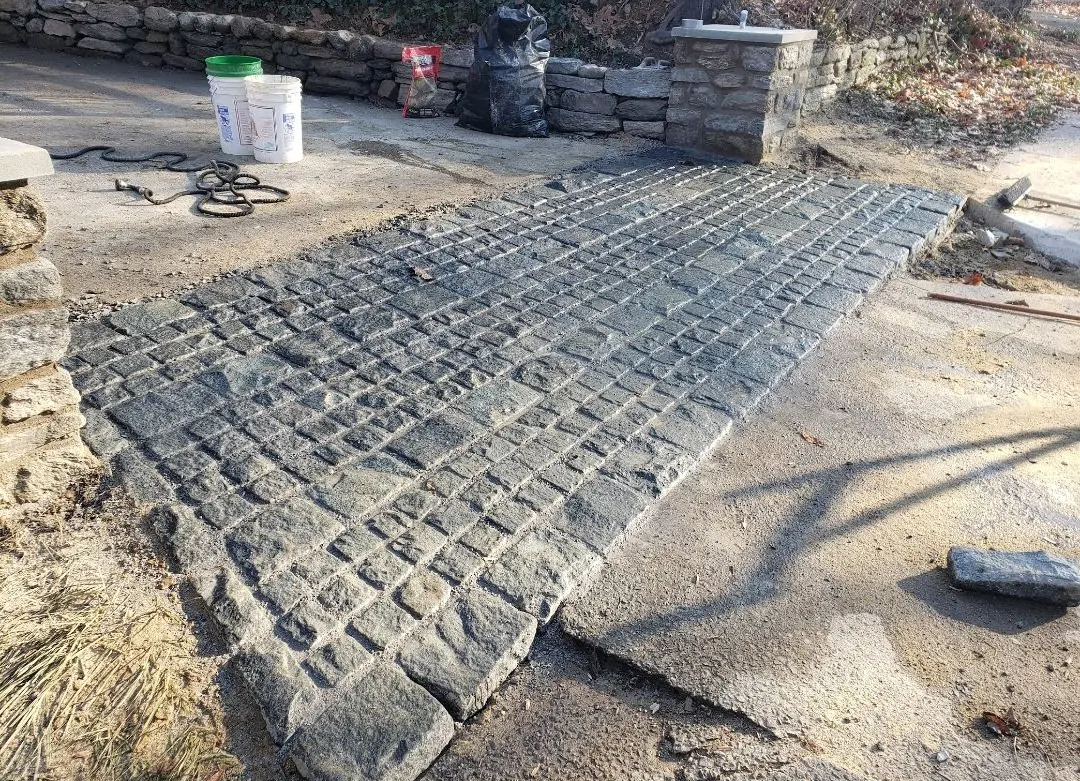 Cobblestone walkway under construction. Stone pillars, cement, buckets, and tools present.