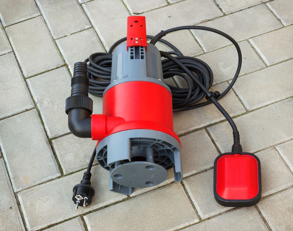 Red and gray submersible water pump with black hose and float switch, on paving stones.