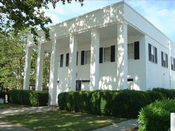 White two-story building with columns and black shutters, surrounded by hedges and trees on a sunny day