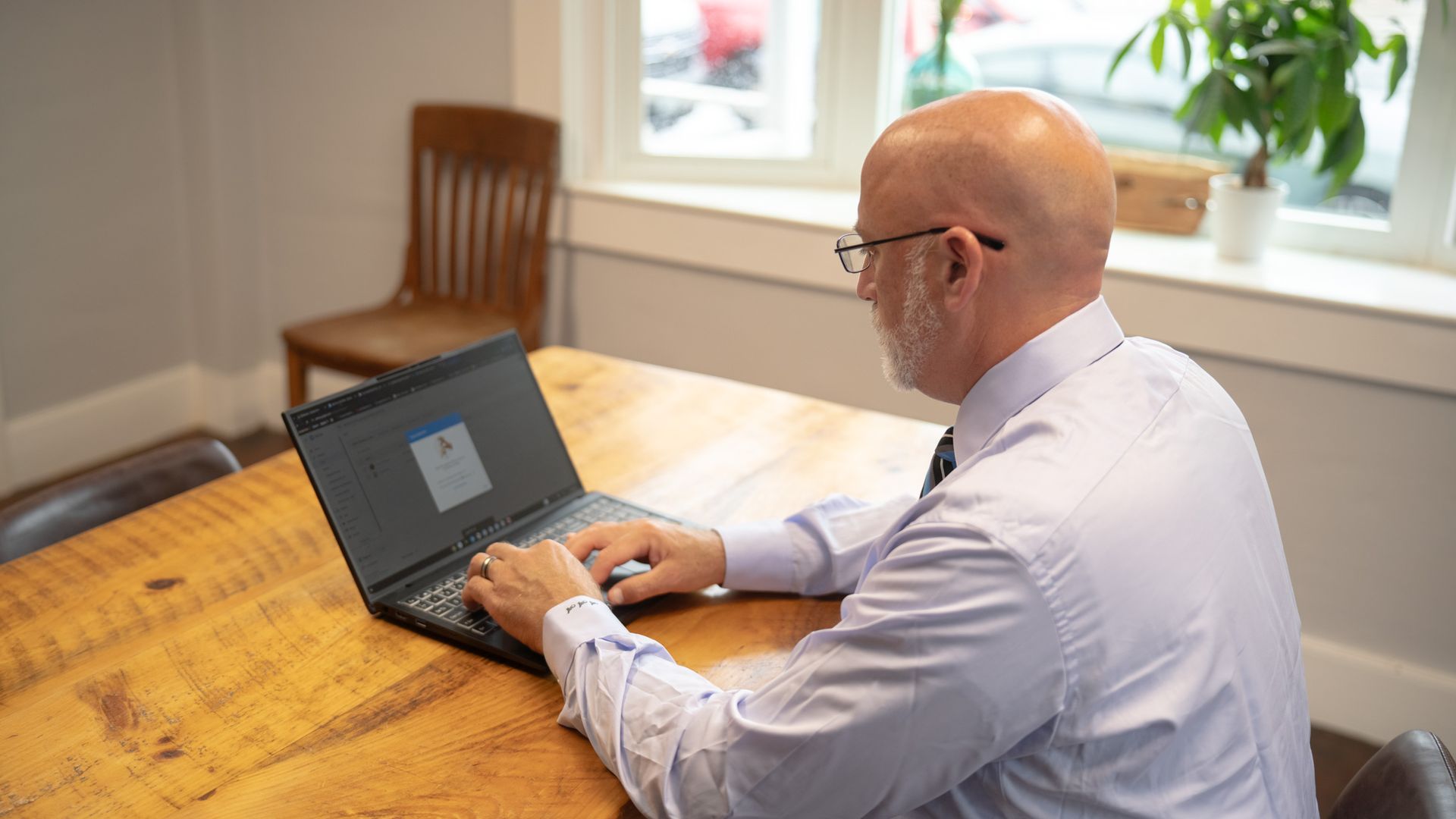 Man typing on a laptop at a wooden table in a bright room with windows and a potted plant.