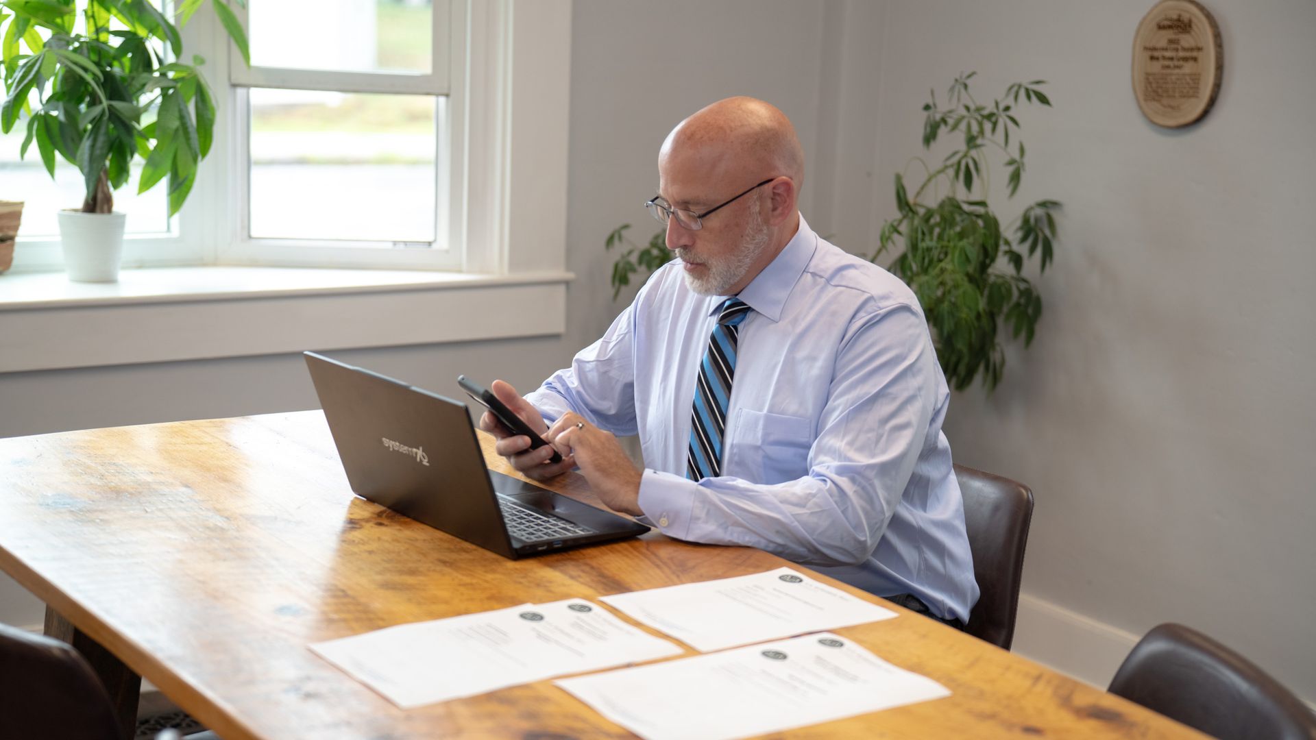Man in a shirt and tie using a laptop and smartphone at a wooden office table with papers and plants nearby