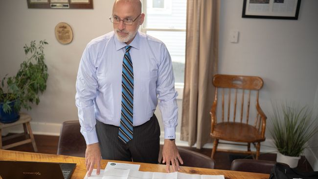 Man in dress shirt and tie standing behind a desk in an office, looking down at papers.