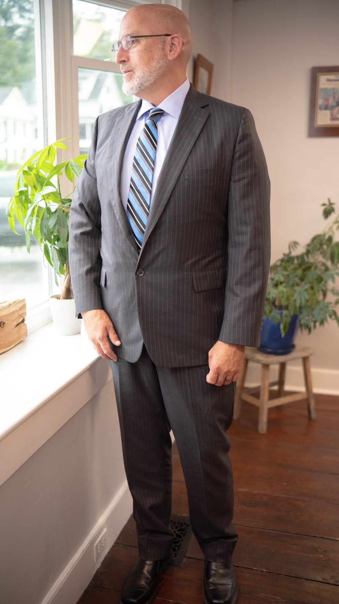 Man in a dark pinstripe suit standing by a window in a bright room with plants.
