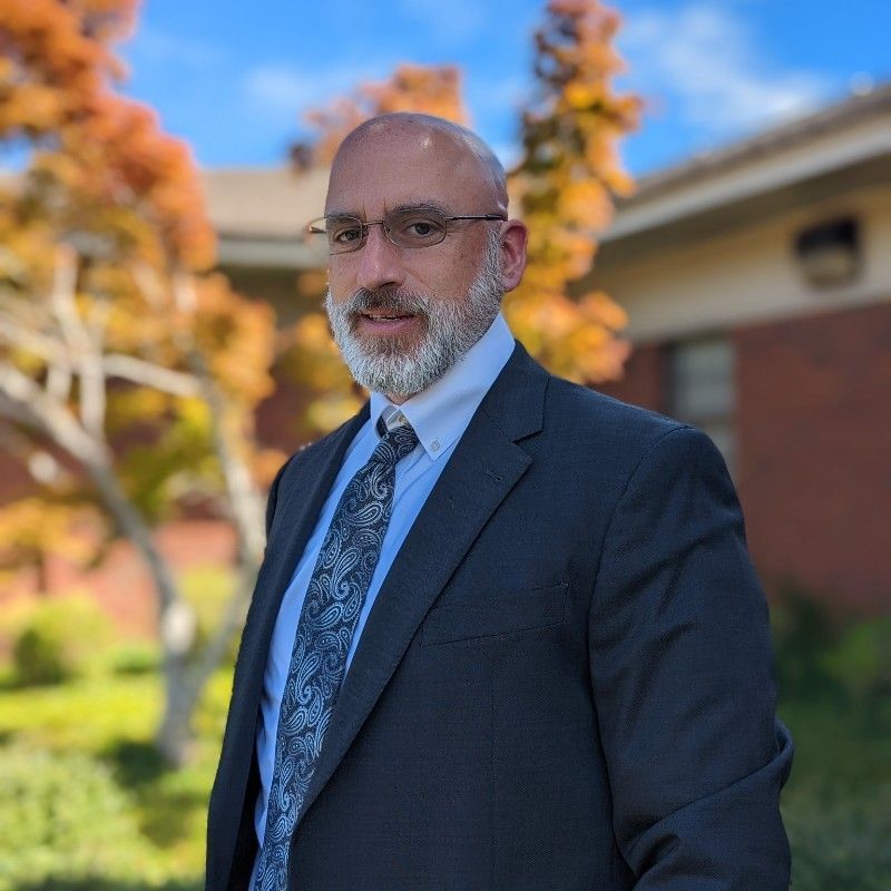Man in navy suit and tie standing outdoors with brick building and autumn trees in the background