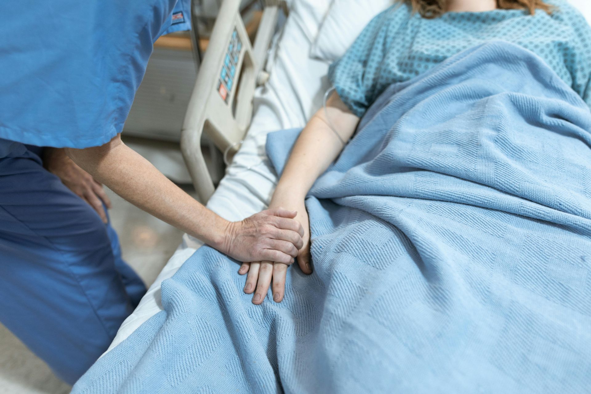 Nurse holding a patient’s hand beside a hospital bed with blue blankets