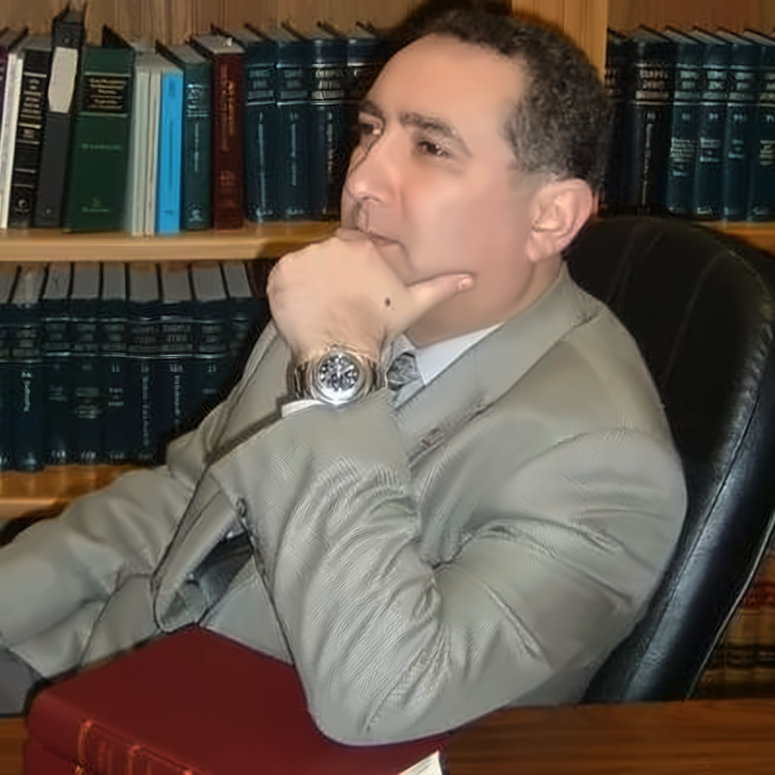 Man seated at a desk in a suit, resting his chin on his hand, with law books on shelves behind him.