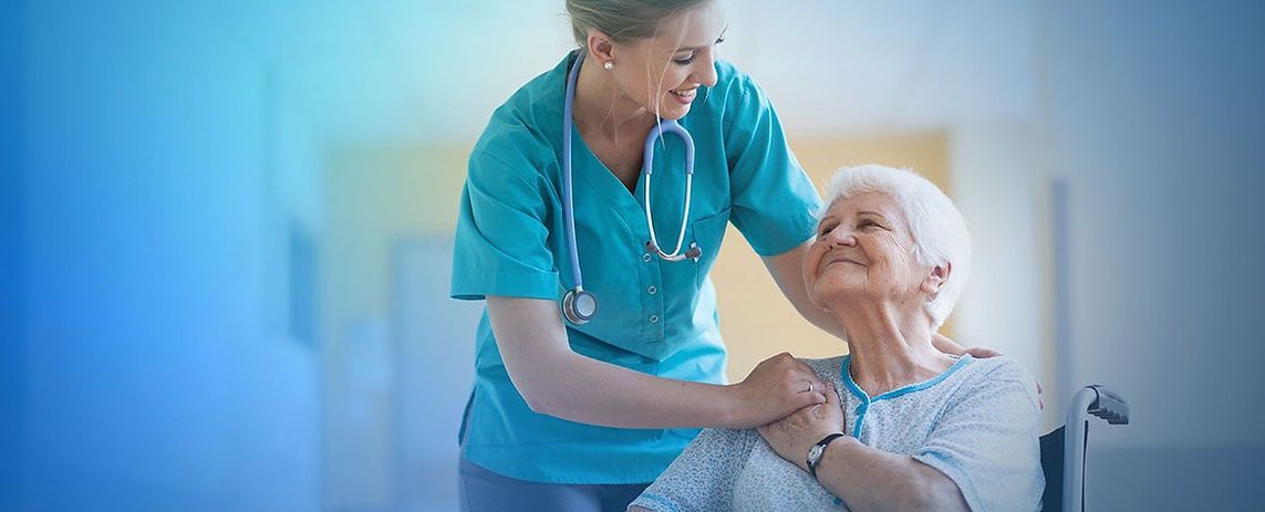 Nurse in scrubs comforts an elderly woman seated in a wheelchair, holding her shoulder and smiling.