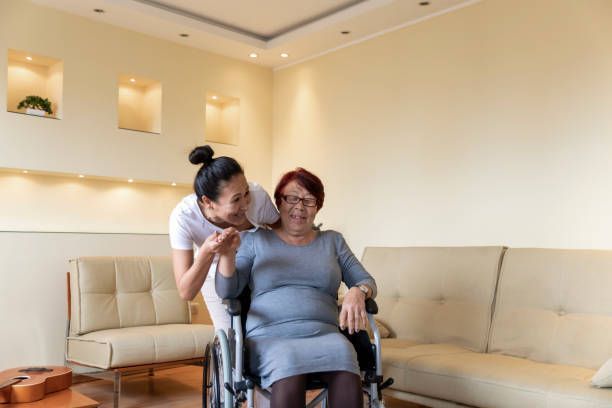 Woman in a wheelchair and caregiver in a living room, smiling. Beige walls and furniture.