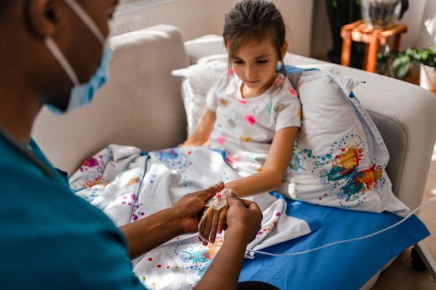 Medical worker tending to a young patient's IV line on a sofa indoors. The girl is seated and looking down.
