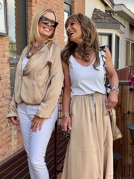 Two women smiling, posing outdoors. One in white pants and gold jacket; the other in a white top and tan skirt.