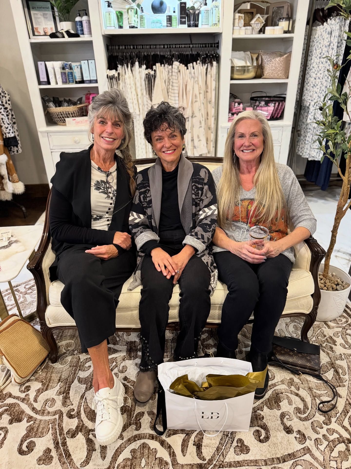 Three women seated on a sofa in a boutique. They are smiling. A gift bag is in front.