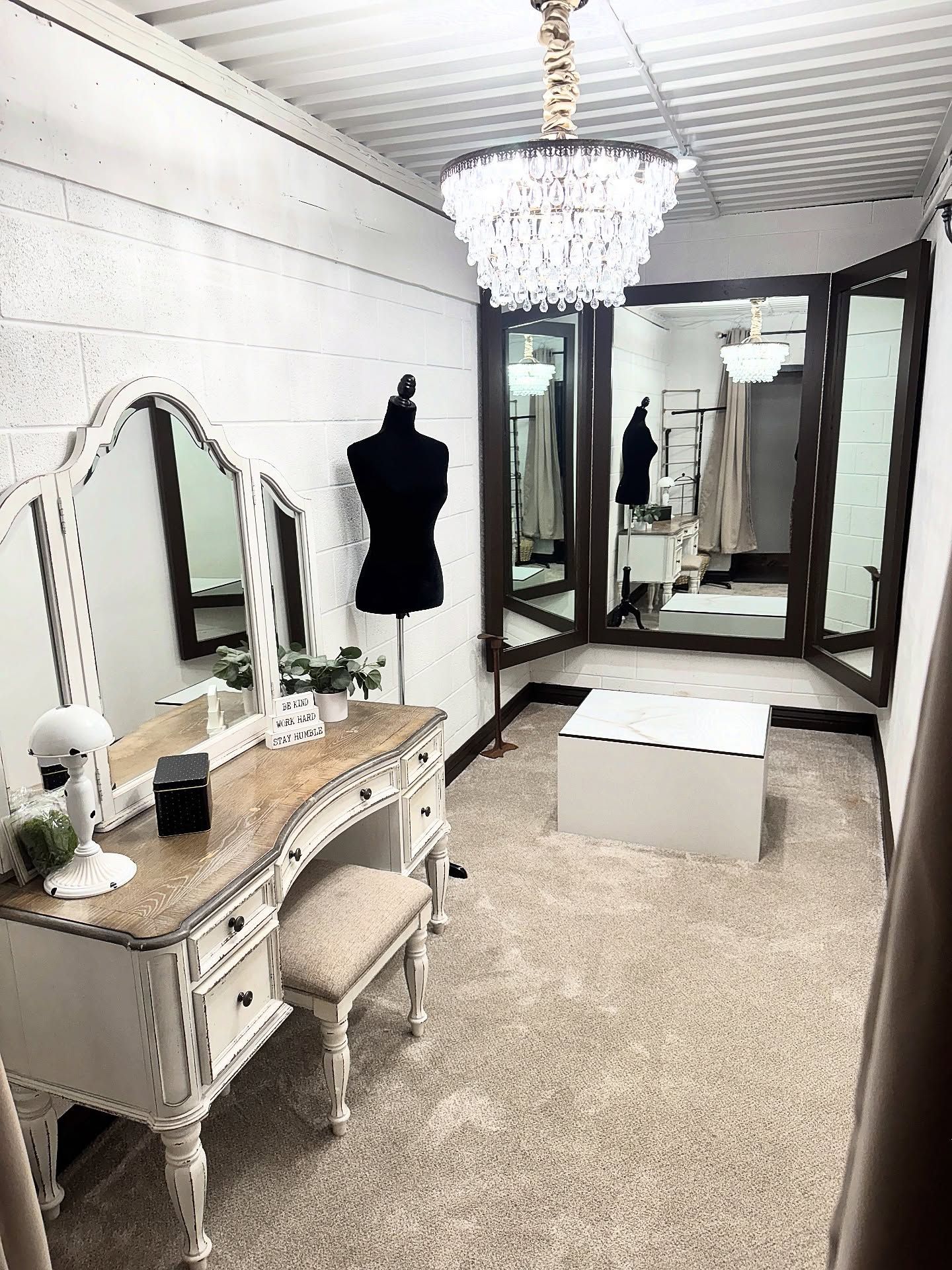 Vanity area in a boutique; white walls, chandelier, dress form, mirrors, and a dressing stool.