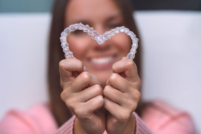 Delle mani che stringono una cornice a forma di cuore tempestata di gioielli davanti a un volto sorridente con uno sfondo rosa sfocato.