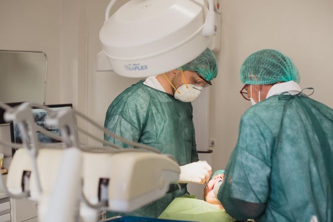 Un dentista con indosso guanti blu tiene in mano una radiografia luminosa di un dente in un ambiente clinico.