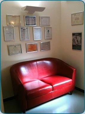 Divanetto rosso in un angolo, sotto certificati e foto incorniciati su pareti beige.