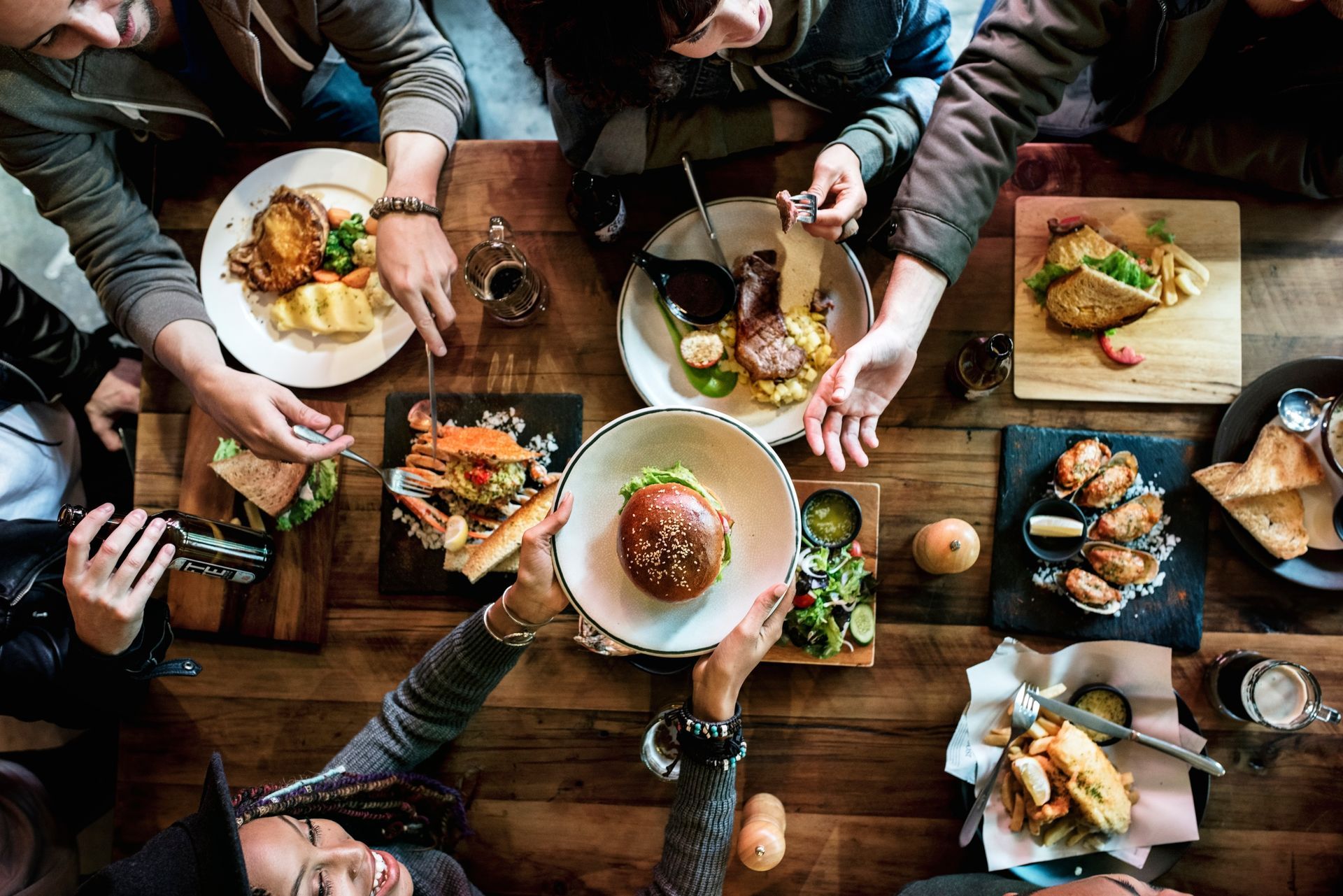 People at a wooden table with various dishes. Hands reaching for food, casual dining setting.