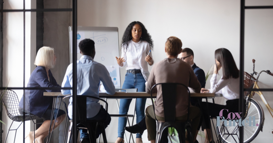 Team meeting in a bright office, one woman standing and speaking to coworkers seated around a table.