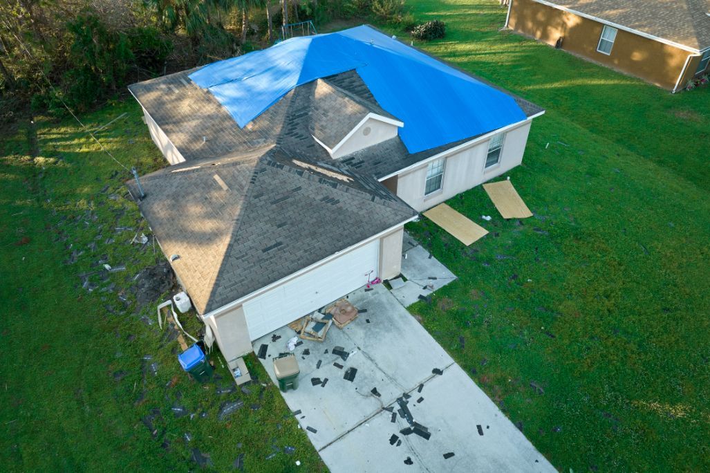 House with blue tarp covering damaged roof, debris on driveway. Green grass surrounds.