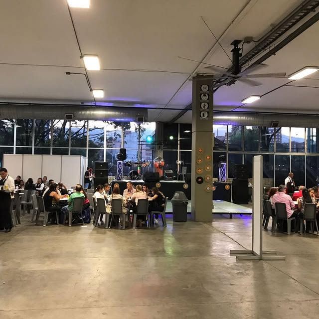 Personas sentadas en mesas en un amplio salón interior con grandes ventanales y suelo de hormigón.