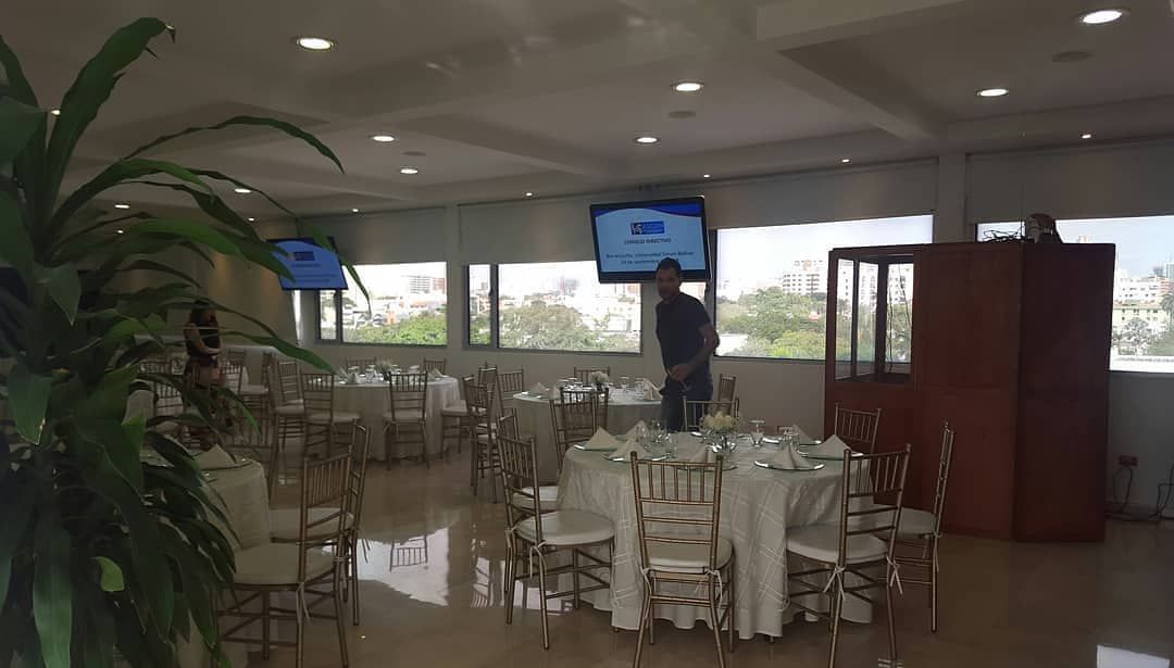 Comedor de restaurante con mesas puestas, una persona cerca del fondo y televisores montados en la pared.