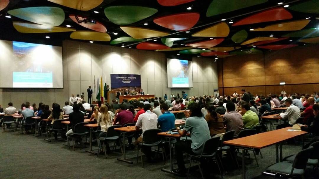 Sala de conferencias con mucha gente sentada en largas mesas frente a un escenario y pantallas de proyección.