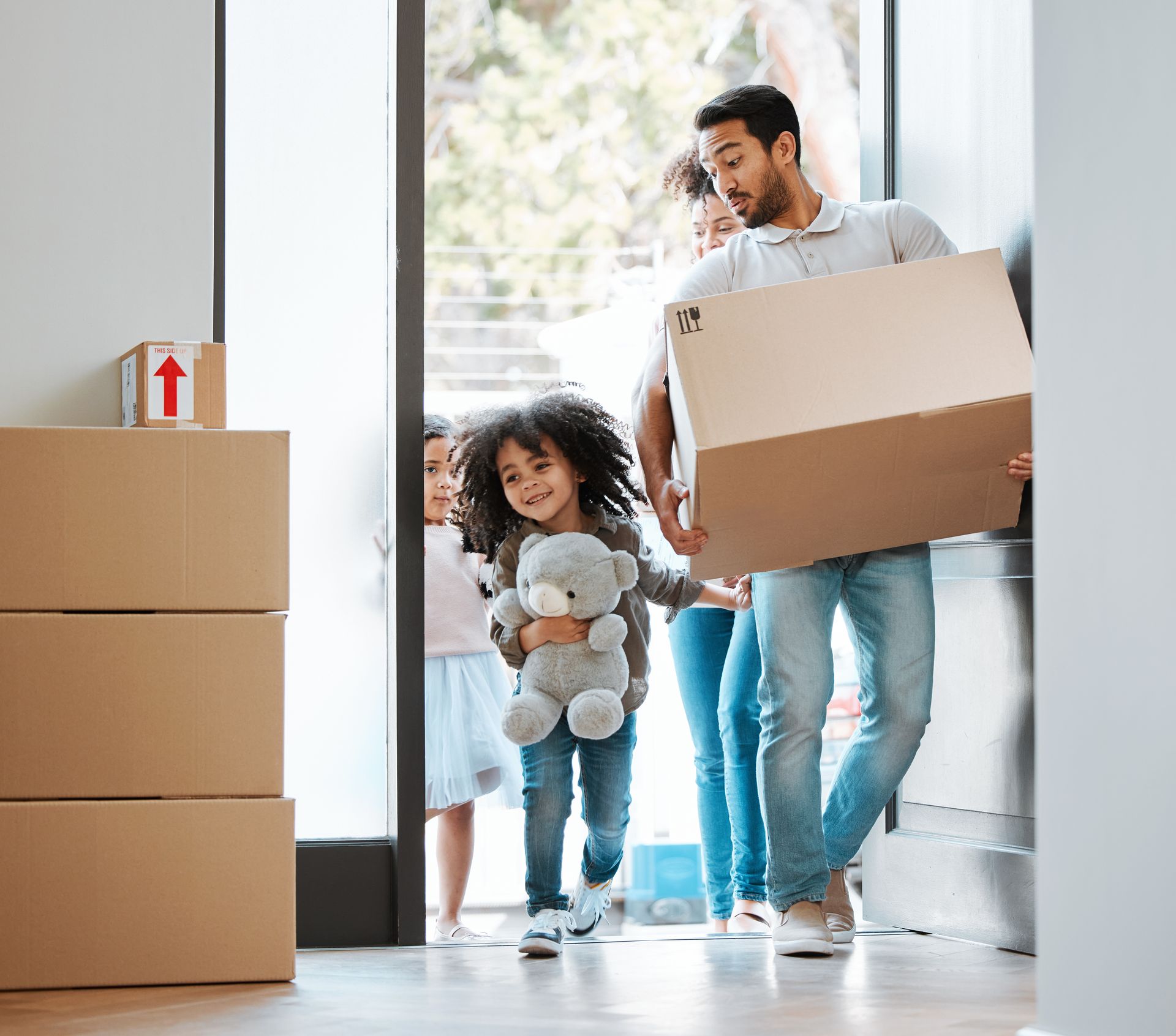 Family carrying boxes into a bright new home, with a child holding a teddy bear.