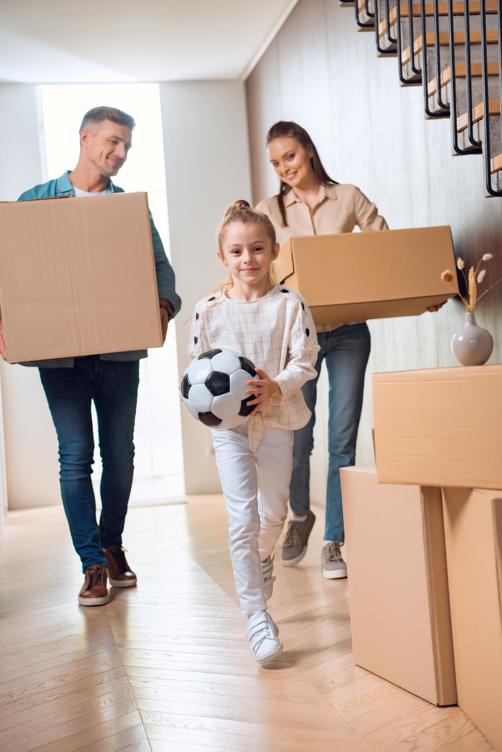 Family carrying boxes into a bright home, with a child holding a soccer ball in front.