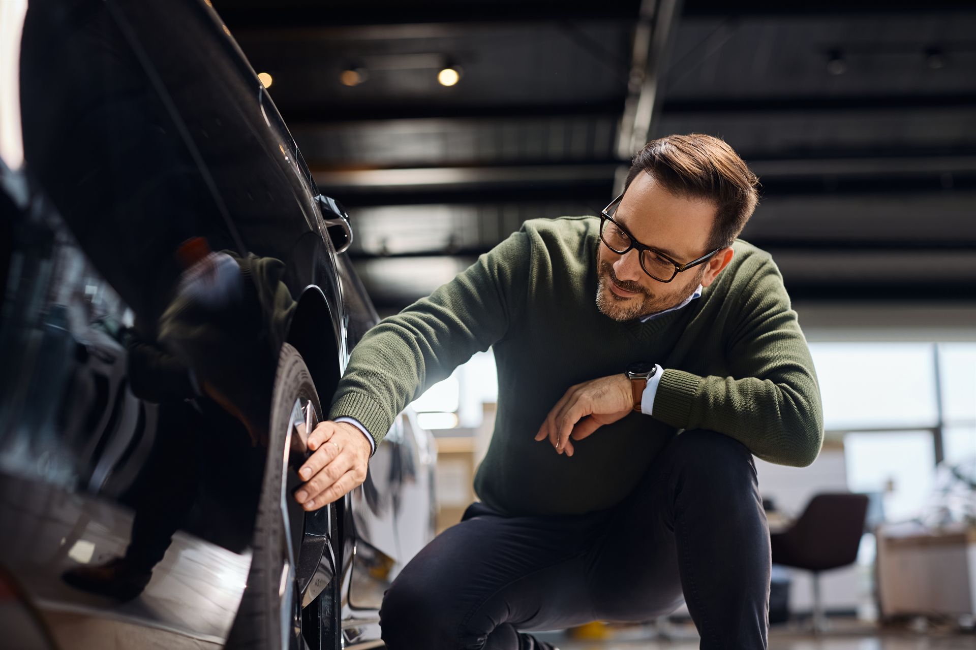 Mechanic inspecting a car tire in a garage, crouched beside a black vehicle.