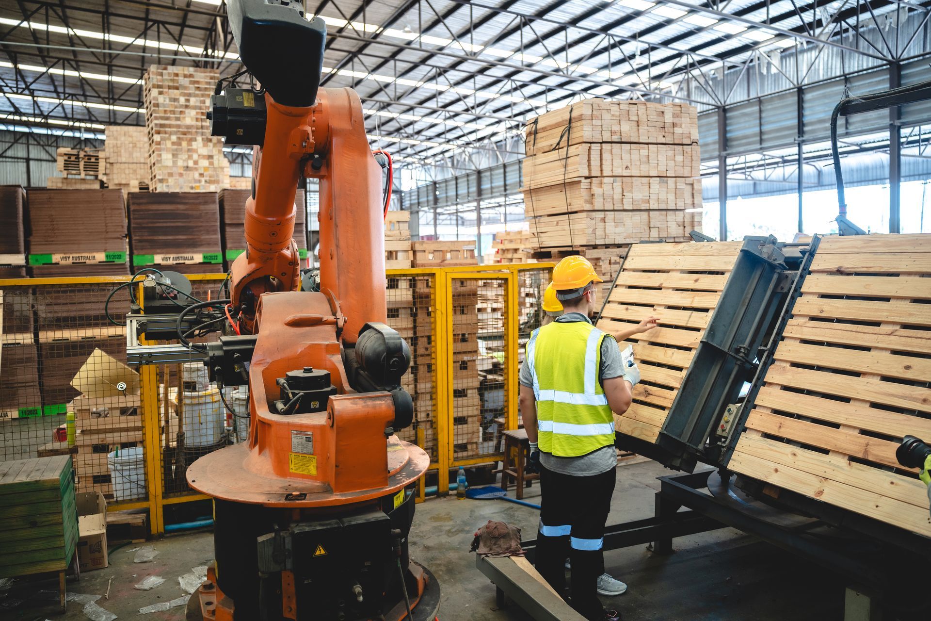 Worker in yellow vest beside orange industrial robot arm in a warehouse, with stacked pallets nearby