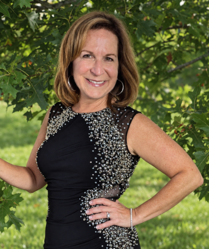 Smiling woman in a black sleeveless dress standing outdoors by a leafy tree