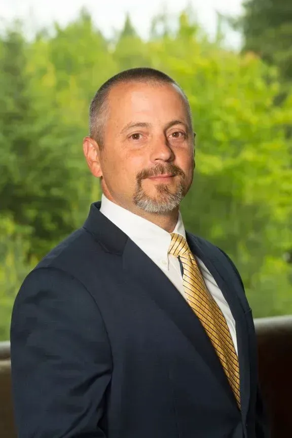 Man in navy suit, gold tie, and goatee, smiling. Set against a blurred green background.